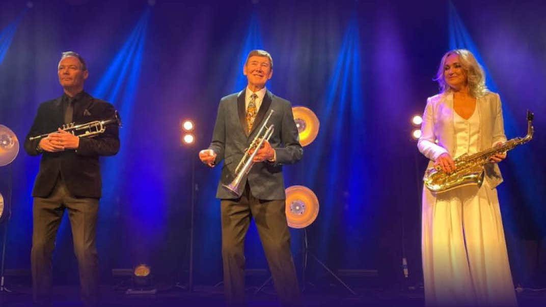 2 men holding brass musical instruments and a woman in a long yellow dress holding a saxophone are spread out in a line on a stage with blue lit background.