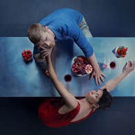 Viewed from above, a man and woman are reaching across a dinner table to each other, looking up at the viewer.
