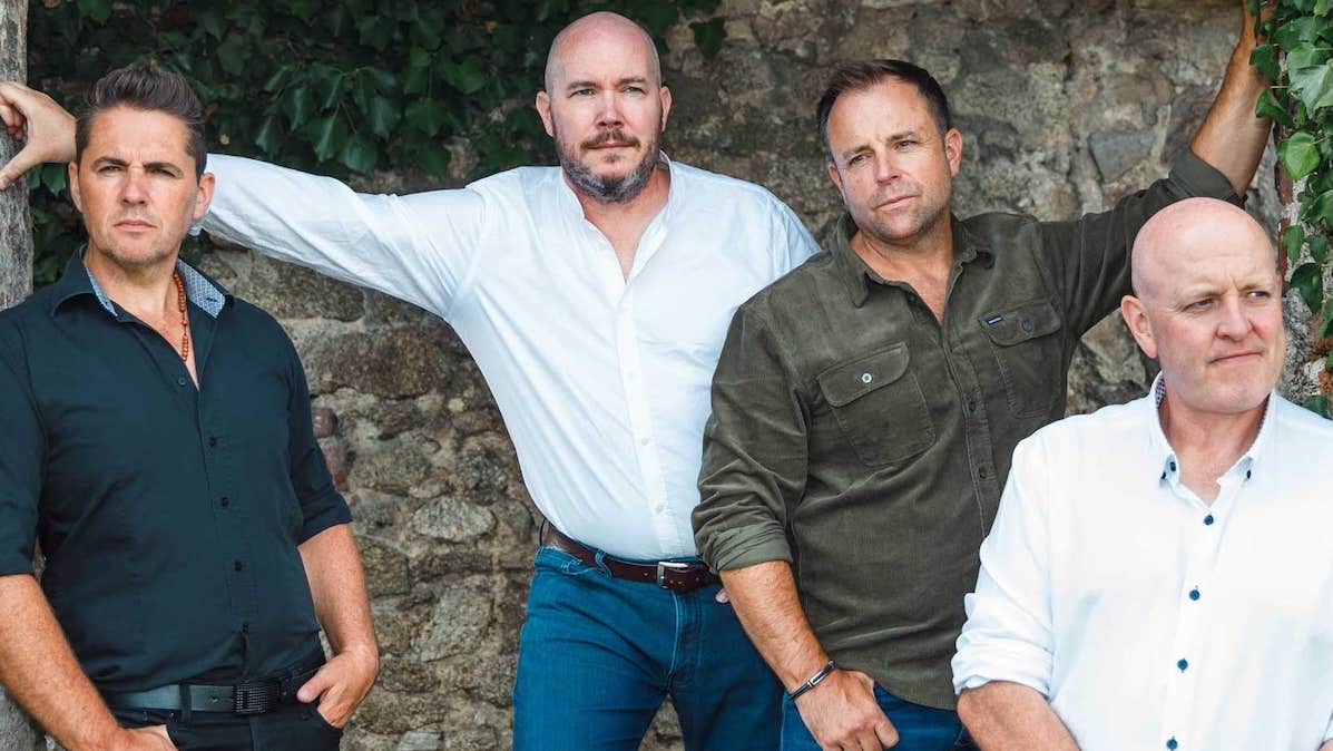 4 men in white or dark shirts are posing against stone building looking away into the distance