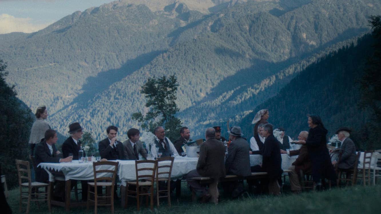A long table outdoors with people seated and empty chairs against backdrop of wooded mountains.