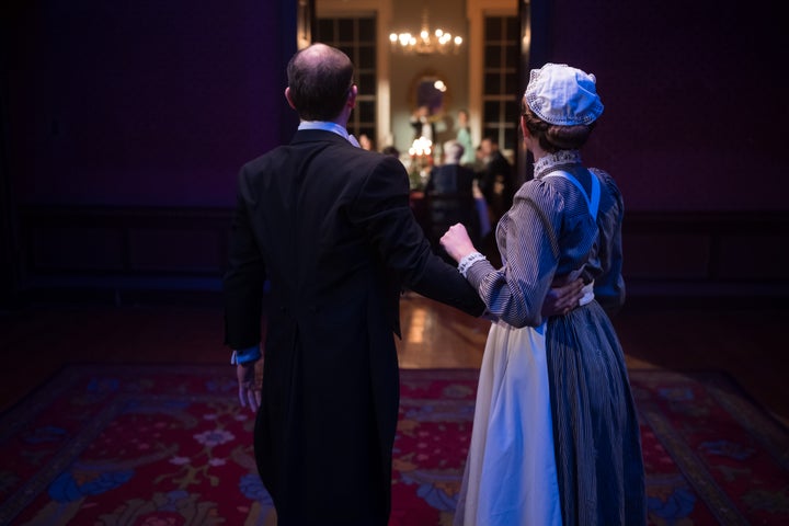 Two characters look into a brightly lit dining room where other characters are.