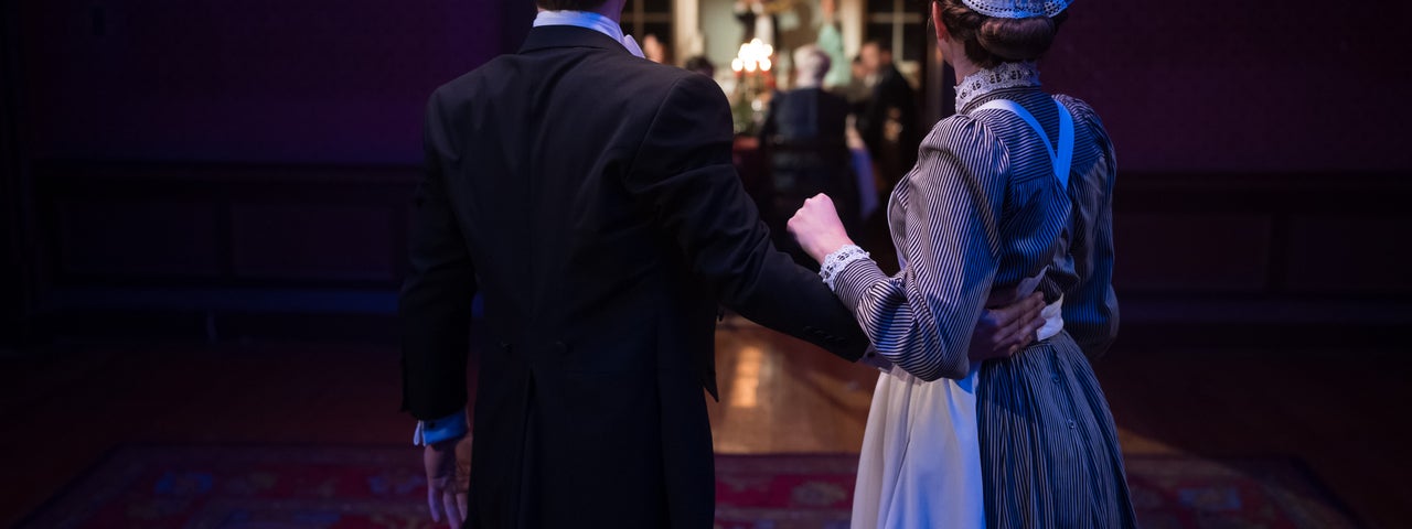 Two characters look into a brightly lit dining room where other characters are.