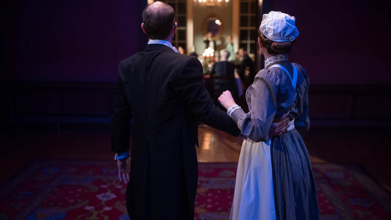 Two characters look into a brightly lit dining room where other characters are.