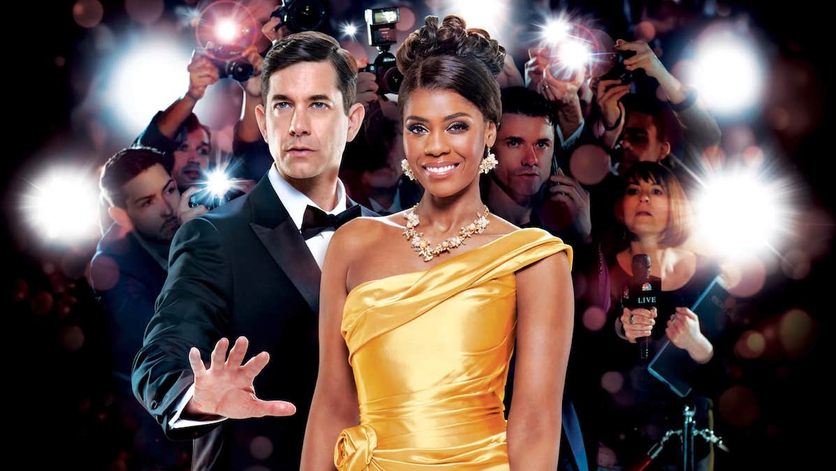 A smiling woman in bright yellow evening gown beside man in tuxedo holding out an open hand in a warning manner