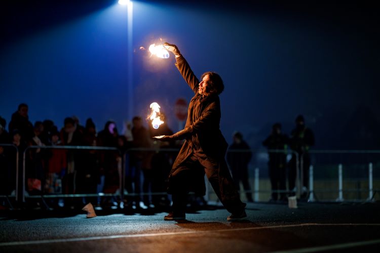 Fire performer lit by the street lamp. While holding fire in each hand, with an audience of onlookers.