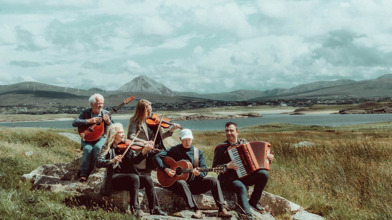 A group of 5 musicians seated on rocks playing with scenic view of mountains and water behind.