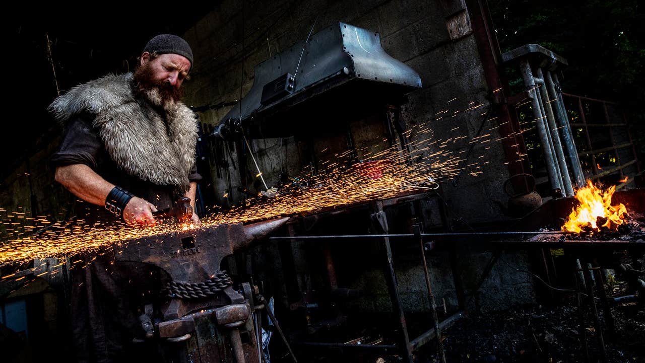 A man in period costume working in a forge with a hammer and anvil