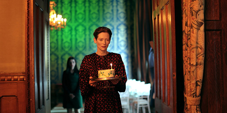 A white woman is walking through a doorway, holding a cake with a single candle. The rooms are high-ceilinged and have dark, clashing colours, making the image feel uneasy.