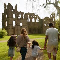 Four people holding hands walking through a garden towards an old stone ruin