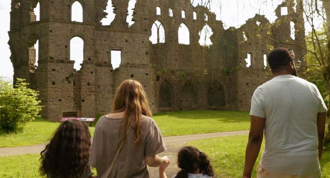 Four people holding hands walking through a garden towards an old stone ruin