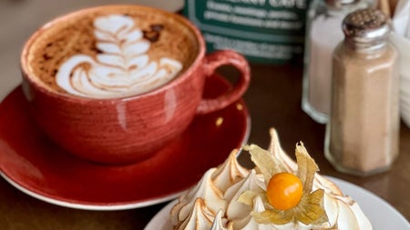 Cappuccino in a brown mug beside a cream tartlet on a white plate