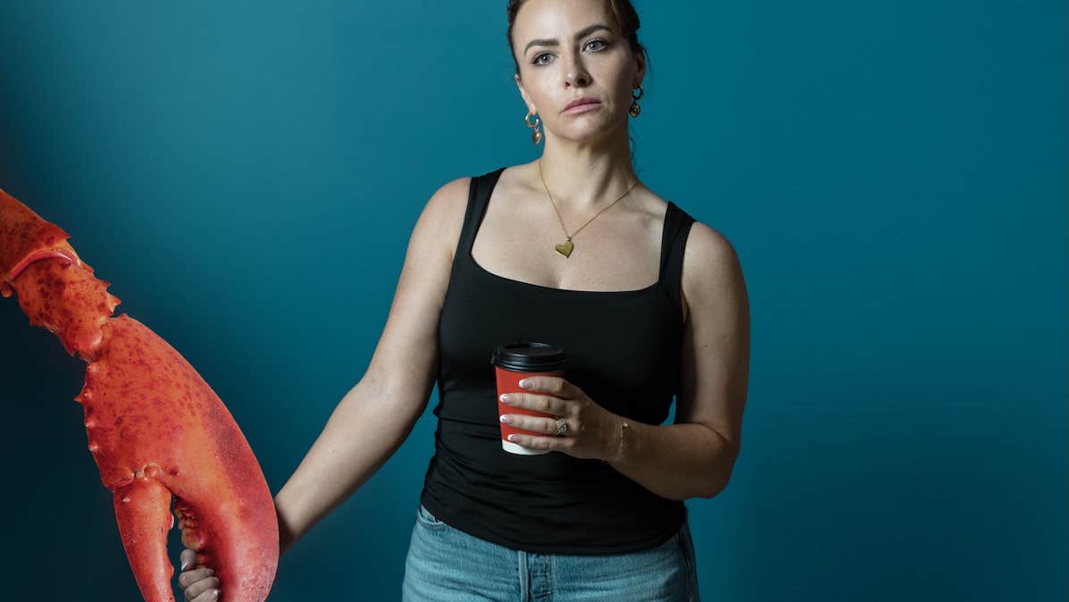Ross and & Rachel by James Fritz, an unhappy looking woman with a takeout cup of coffee is holding the large claw of a large lobster.