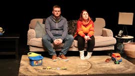 On a stage, 2 people are seated on a sofa with children's toys on a mat in front of them.