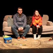 On a stage, 2 people are seated on a sofa with children's toys on a mat in front of them.