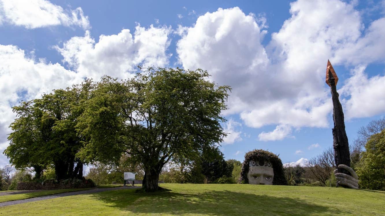 Green field with a giant head poking up from the ground holding a very tall spear