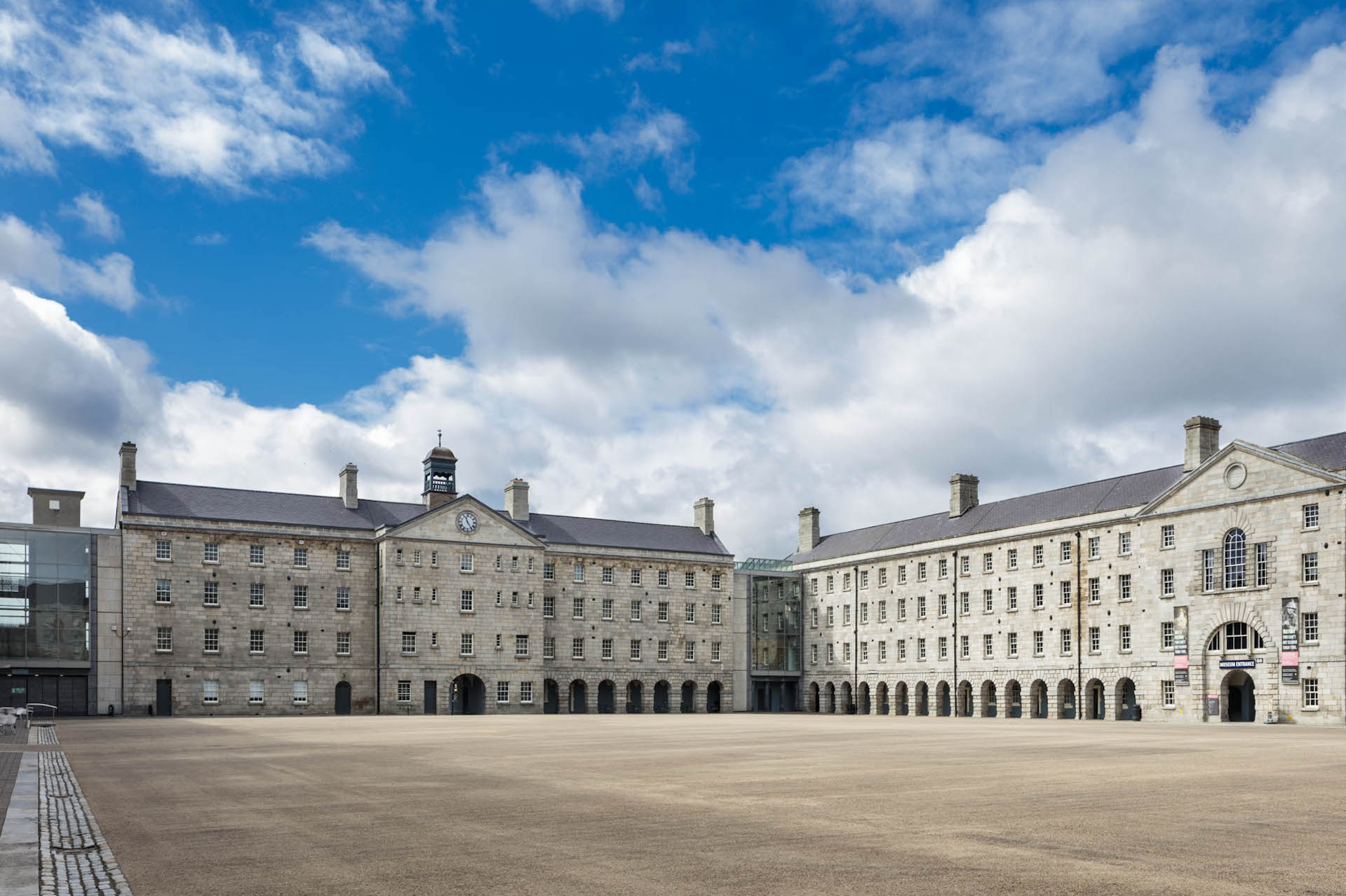 Large four story buildings with lots of windows and arches at ground level, at right angles forming a large square in the middle with blue cloudy sky above.