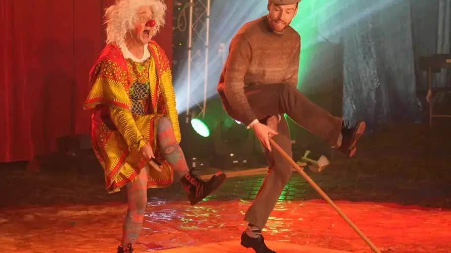 2 people dancing on a stage, one dressed in patterned clothes wearing a white wig and red nose.