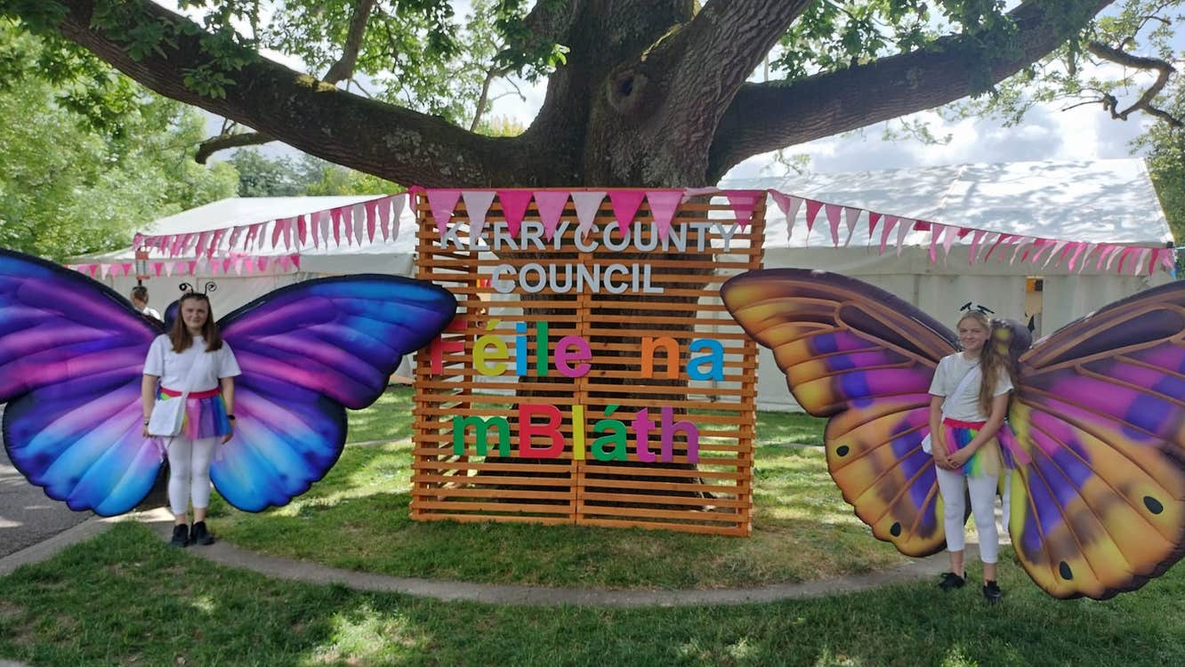 Name of event in coloured letters on a display under a large tree, standing either side are 2 people dressed in white with large, multicoloured butterfly wings.