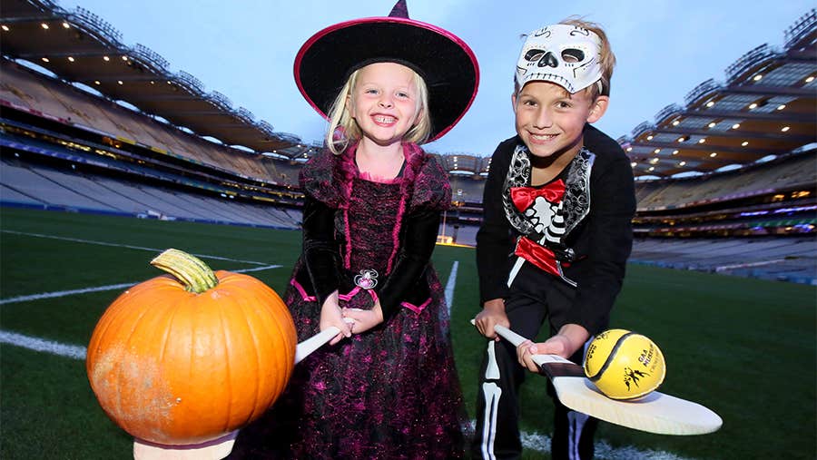 2 smiling young children in Halloween costumes, holding out hurlies with pumpkin on and view of large stadium in the background.