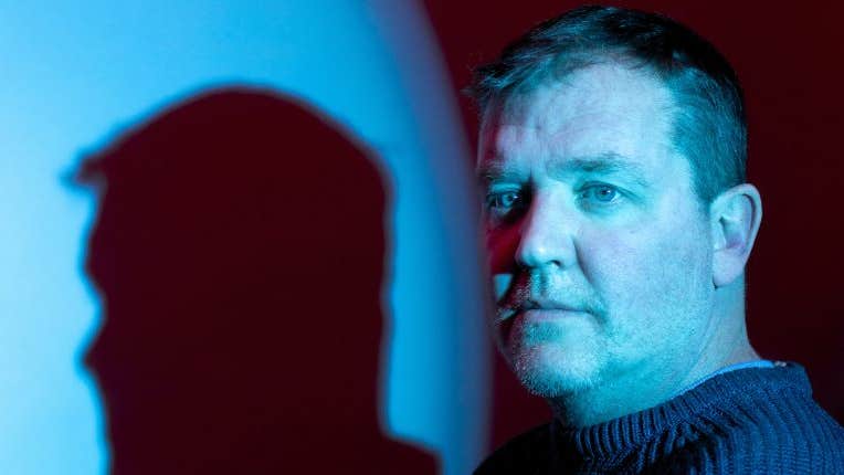 A serious looking man lit in blue with shade of his head cast to one side of him.