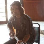 A serious looking man seated on a chair with a guitar with back to the light from a doorway