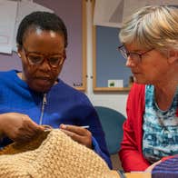 A woman knitting with another woman looking on seated beside her at a table