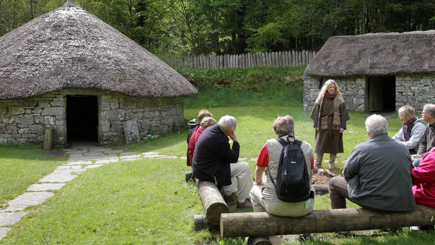 a group of people sitting on log seats in front of thatched round huts while a tour guide in ancient dress speaks