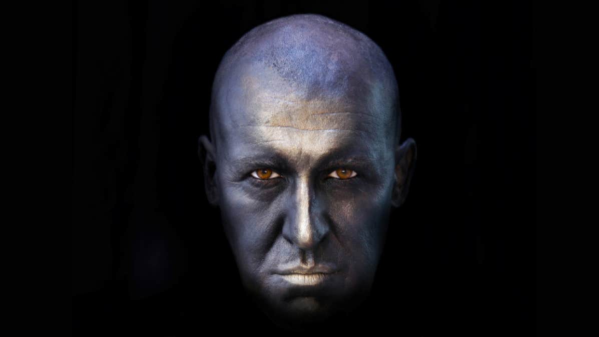 UNDERNEATH written and performed by Pat Kinevane - a bald man's face in silver colour surrounded by blackness.