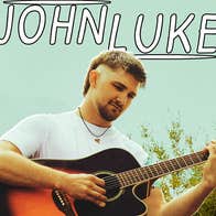 Singer/songwriter and musician Johnluke, a slightly smiling man is seated on a rock playing a guitar, surrounded by greenery