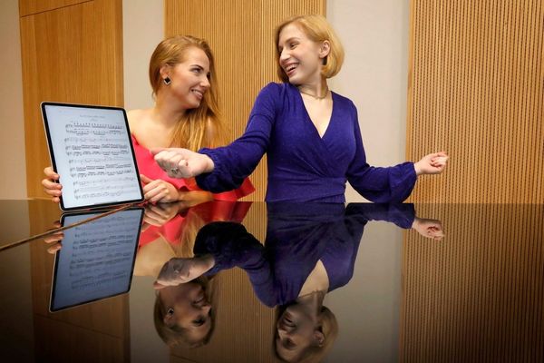 2 women standing behind a shiny surface with a music score, laughing looking at each other .