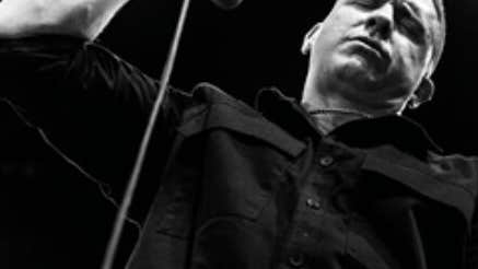 Black and white image looking up at a serious looking man, holding a mic.