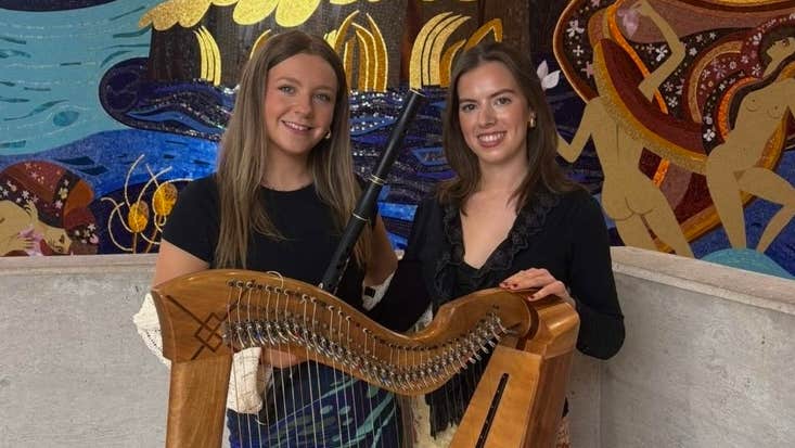 Harper and fiddle player Aoibhe Kettle standing with another young woman behind a large, wooden harp.