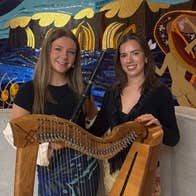 Harper and fiddle player Aoibhe Kettle standing with another young woman behind a large, wooden harp.