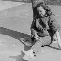 Black and white photo from 1958 of a woman sitting on the ground at the base of a sculpture, looking down at her lap.