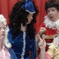 4 people in wigs and costumes are talking with red curtains behind them.