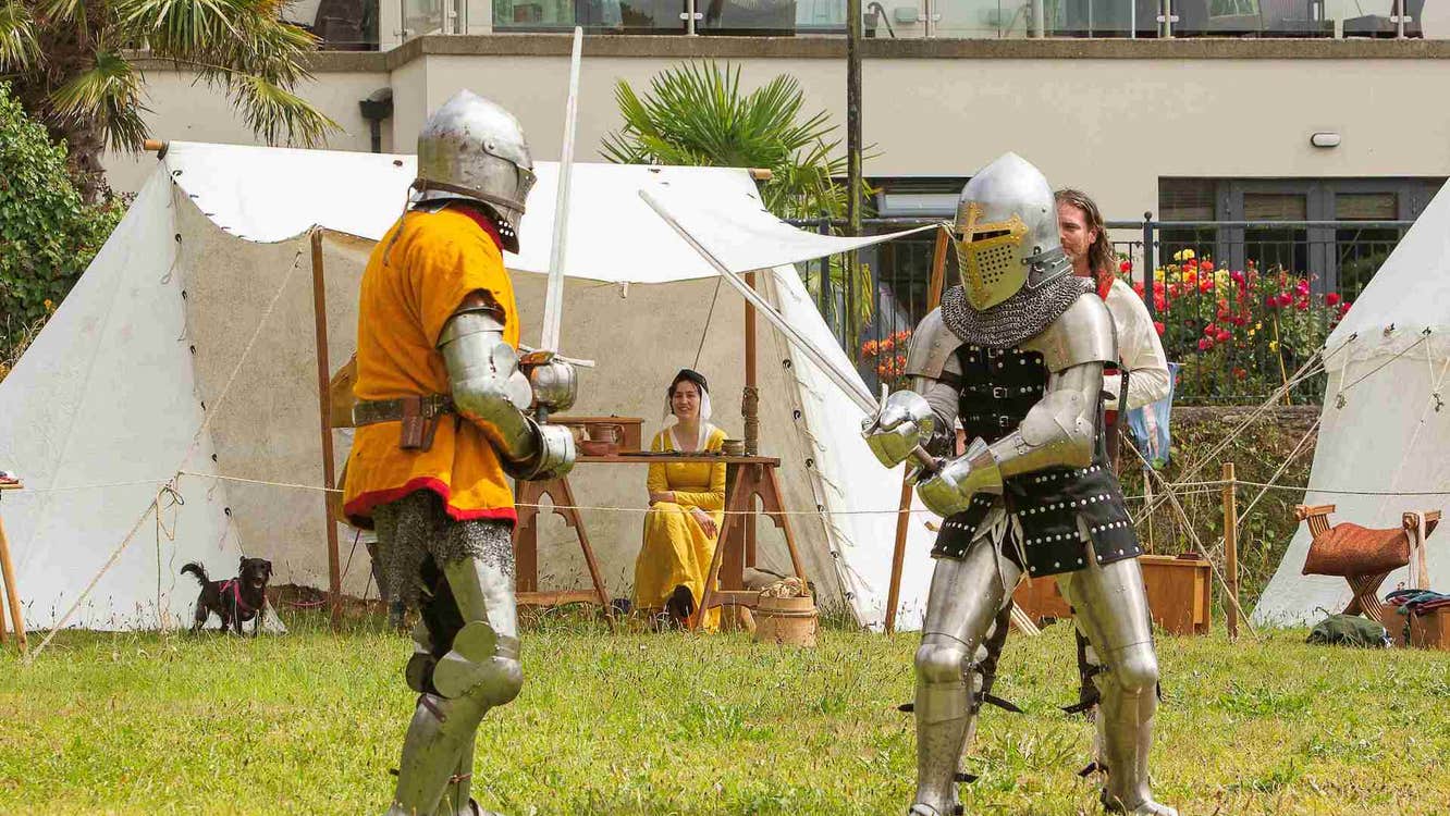 Battle reenactment - fun on the field, 2 people dressed in suits of armour and chainmail with swords, white tent in the background.