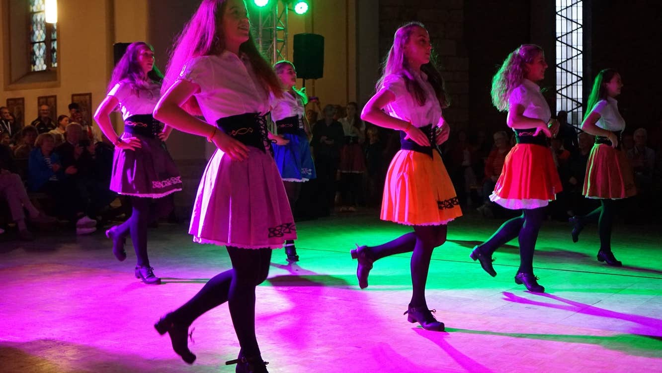 Group of 6 irish dancers spread out in 2 lines in hard shows with audience looking on.
