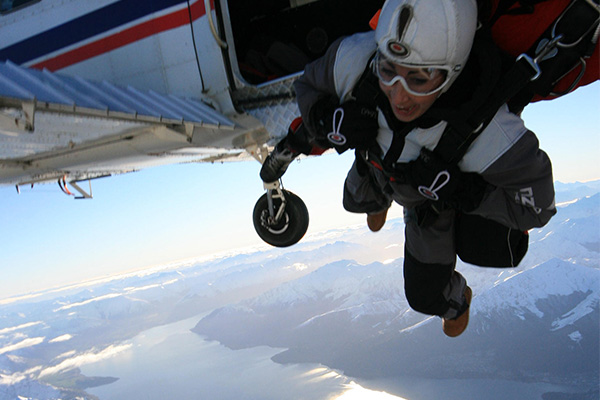 FREEFALLING by Georgina Miller. A person dressed in jumpsuit with helmet and goggles just outside a small plane, flying about some snow capped mountains.