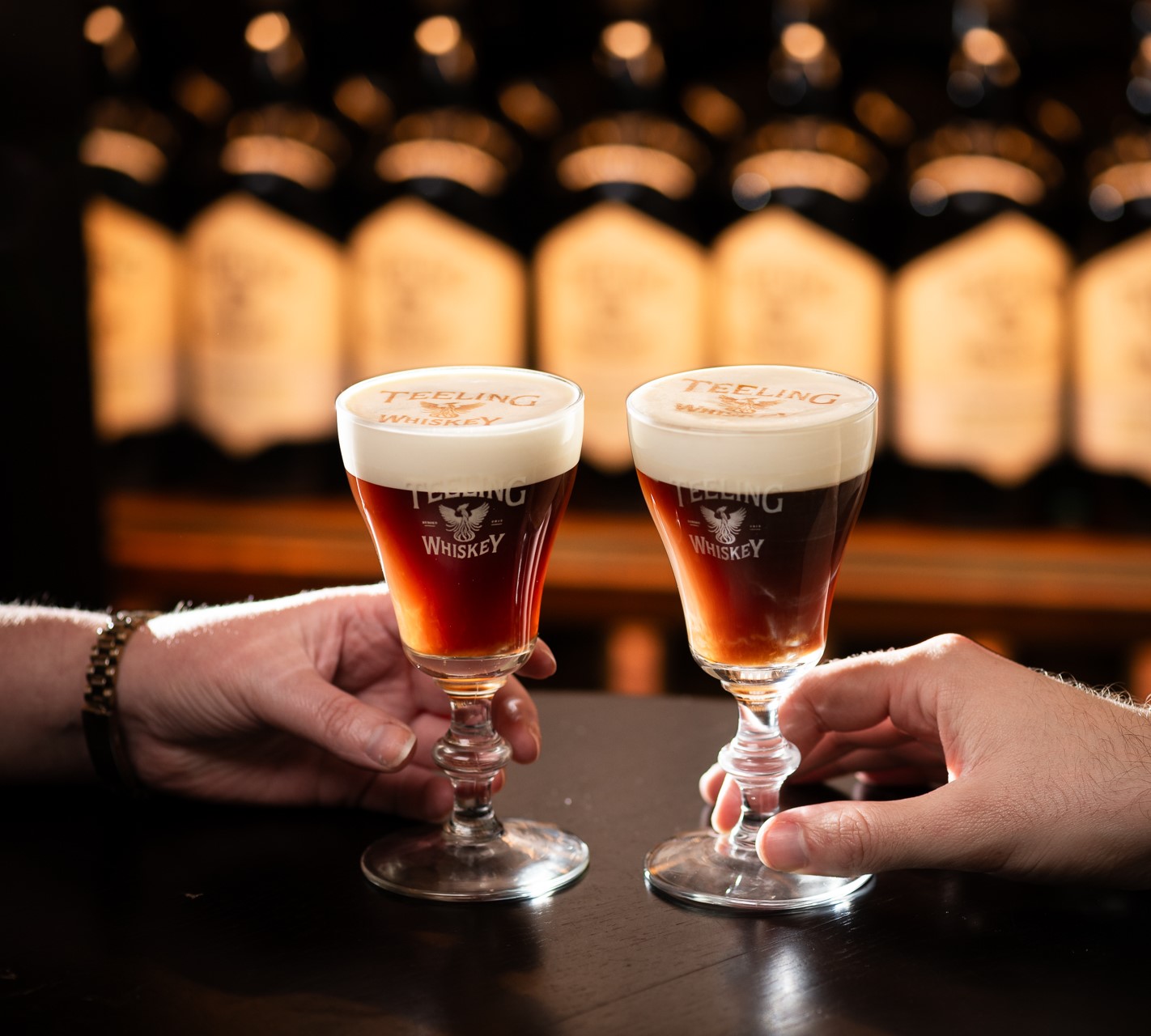 Two goblet glasses containing brown drink with white creamy top, a hand holding the bottom of each glass on a wooden counter top.