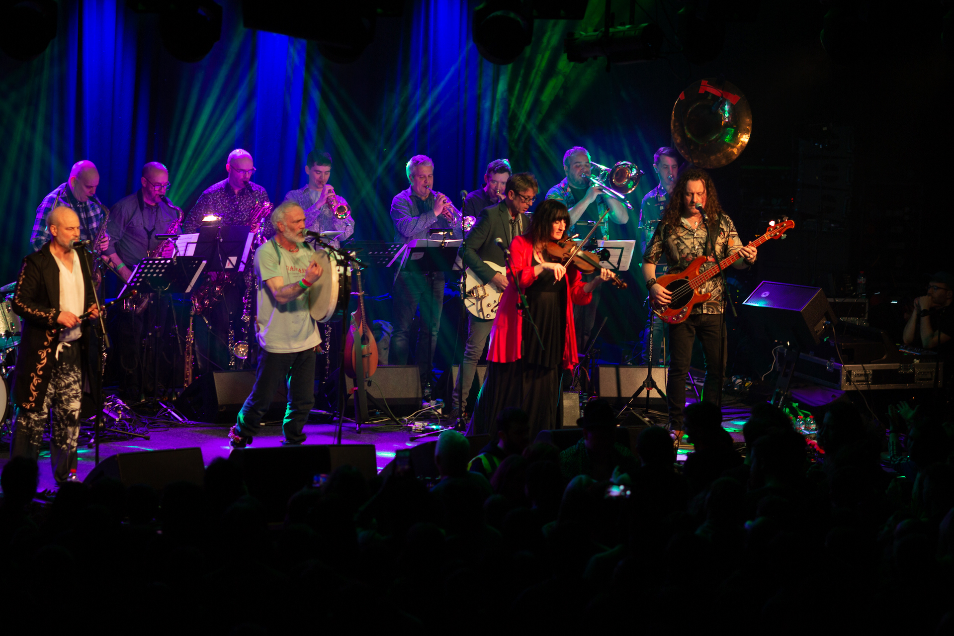 On a stage, a line of musicians at the back playing brass and wind instruments, in front more musicians playing guitars, fiddle.