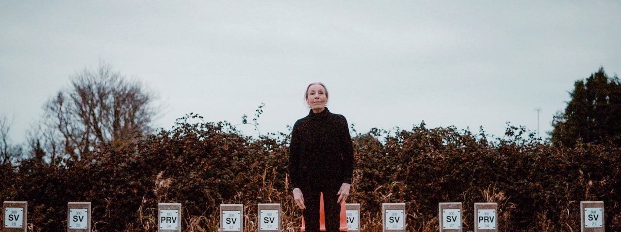 An older woman dressed in black is standing outdoors on a dull day on concrete area with 2 lit fluorescent tubes on the ground either side of her
