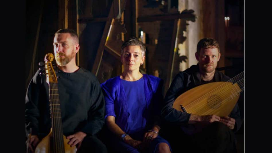 3 people looking serious sat in a line holding musical instruments, dimly lit.