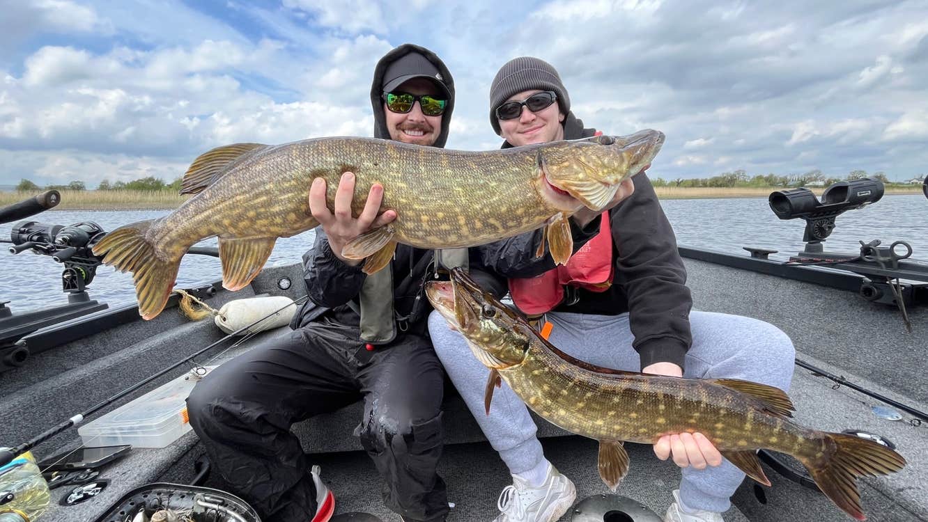 Two anglers holding large pike aboard a fishing boat on a lake
