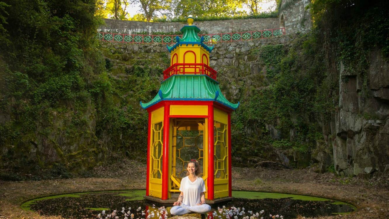 A small yellow and red round pagoda shaped building with green roof surrounded by tall, green covered walls with a person seated in front in a yoga position.