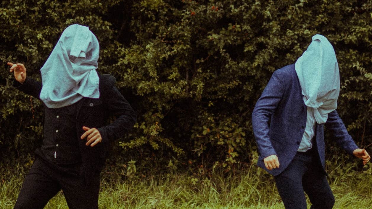 2 people outdoors with hedge in background, standing apart with large, pale blue garments over their heads.