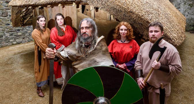 People dressed at Vikings holding weapons and a green and black shield outside a thatched replica Viking house