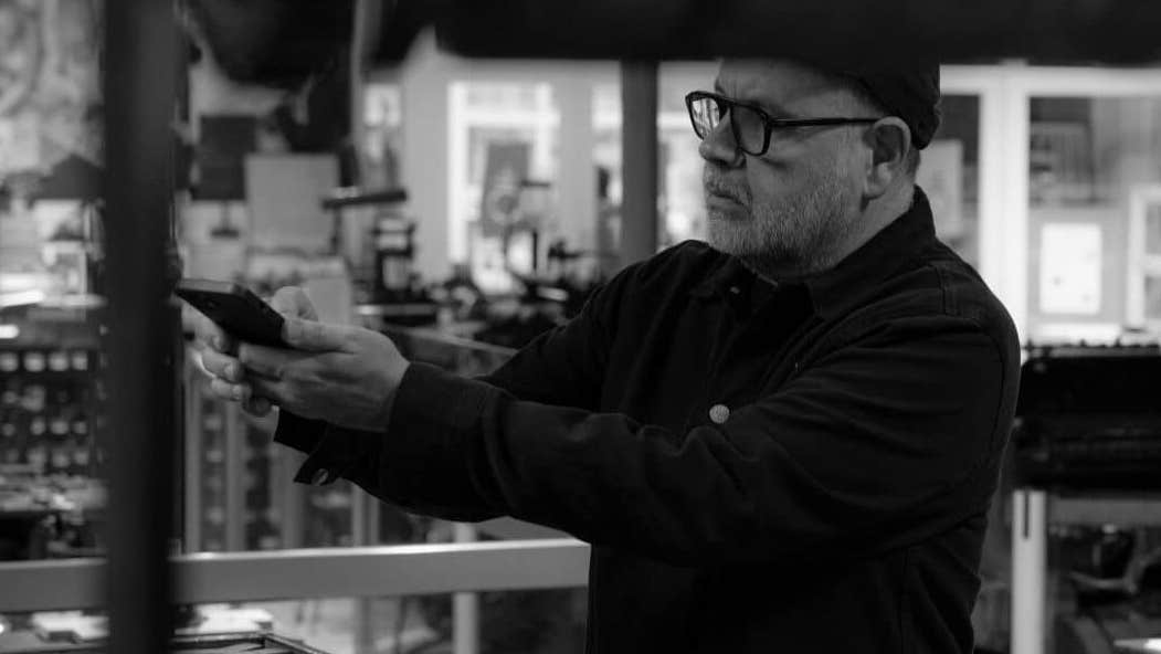 Anthony Burrill and Maser In Conversation, a man in a museum holding his mobile phone horizontally out in front of him with various print machines in the background.
