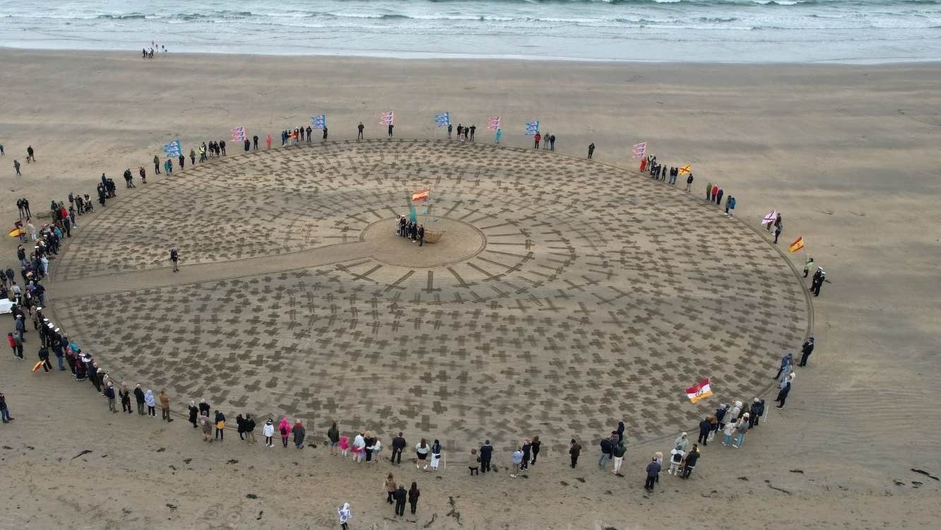 People form a circle around crosses marked in the sand at a beach memorial event