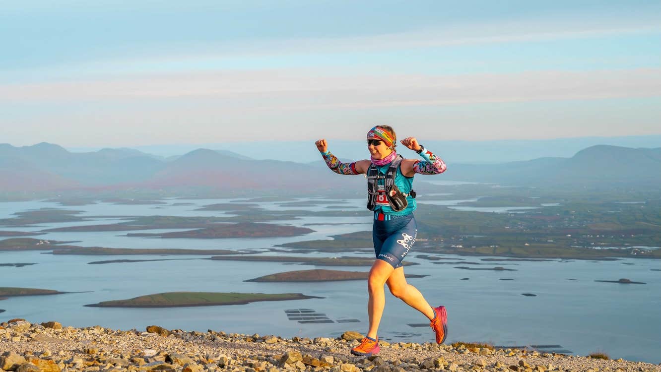 Westport Sea 2 Summit Adventure Race - a person running with arms raised, on top of a high hill with water and hills below in the background.