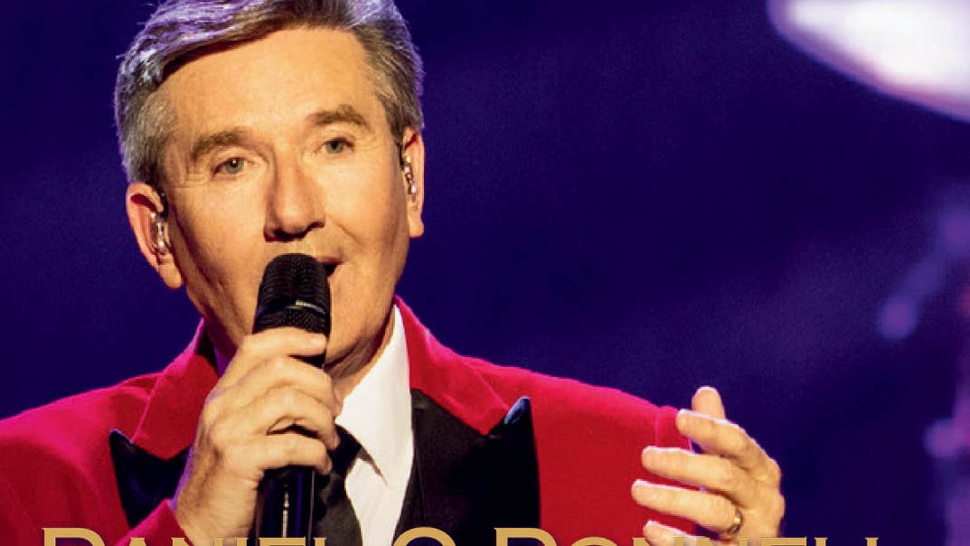 A man in red jacket and black tie is singing into hand held mic against dark purple background.